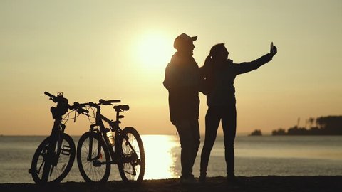 Silhouettes of two cyclists at sunset. The pair finished the bike ride on the river and do selfi on a beautiful background. Couple in love. Unusual date. hotograph on a mobile phone
