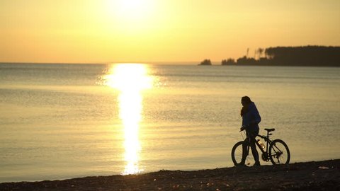 Girl with a bicycle goes along the beach at sunset. Silhouette of the girl at sunset. Loneliness. Thoughtful girl. Romantic girl. Silhouette of the girl: looking into the distance