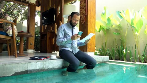 Businessman working with smartphone and documents by pool in outdoor villa
