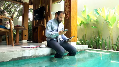 Businessman working with smartphone and tablet by pool in outdoor villa
