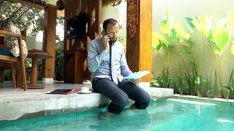 Angry businessman with documents talking on cellphone sitting by pool in outdoor villa

