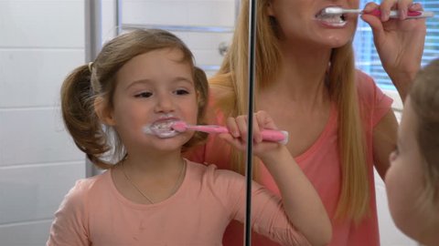 Young attractive mom and her cute daughter brush their teeth in front of the mirror.