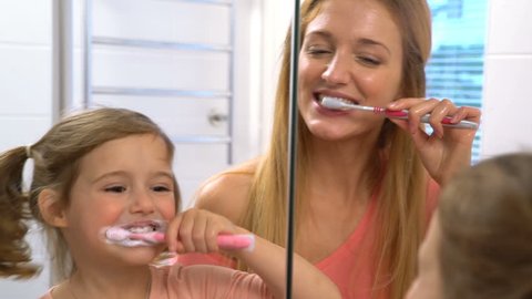 Young attractive mom and her cute daughter brush their teeth in front of the mirror. Slow motion.