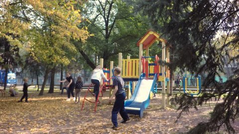 Late fall. In the background, drive cars.  Young children having fun. Next to the children's playground are growing spruce. A man sits on a bench in the park. Little boy playing with his mother. 