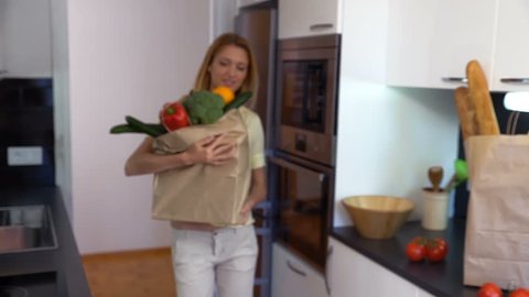 Happy young woman brings to the kitchen a large paper bag of groceries and smiling at the camera. Slow motion.