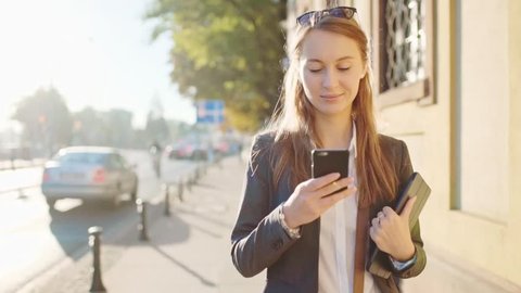 Young Businesswoman Using SmartPhone, Going to Work in the Sunny Morning City. SLOW MOTION. STEADICAM Stabilized Shot. Attractive Professional Business Woman with a cell phone in a city. Lens Flare.