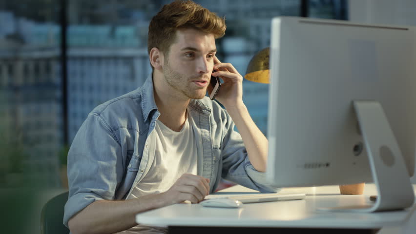 4K Young man using computer & talking on cell phone in city apartment. Shot on RED Epic.