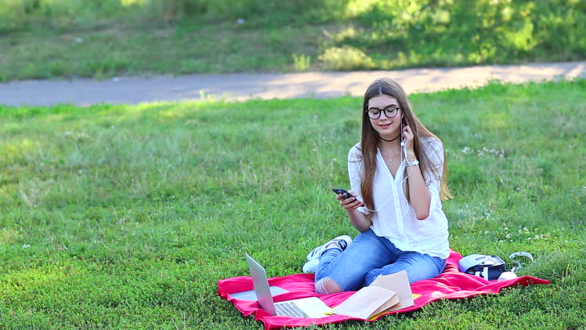 Young Beautiful Female With Long Hair and Glases in Jeans and Shirt Smiling and Enjoying the Laptop. a Woman Sitting in the Park, Doing the Work or Studying, Doing Homework. Lady Sitting on a