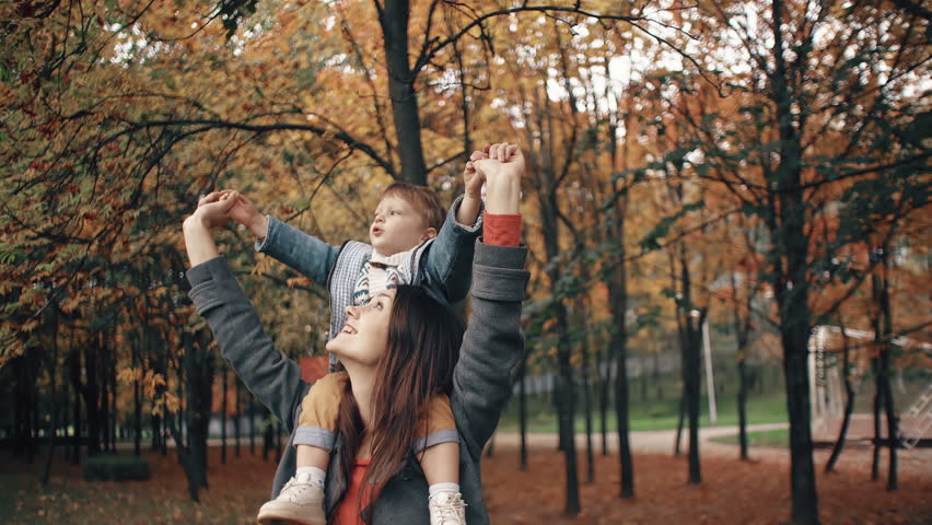 beautiful young mother holds on the shoulders a cute little son, the boy point the finger at something in autumn park 4k