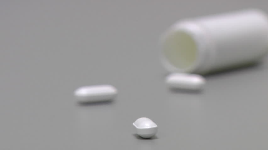 One pill in focus, two pills and white container out of focus.