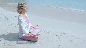 Healthy mature woman sitting in yoga position in a sunny beach destination, meditating holiday outdoors. Sport recreation exercising training, aspirational lifestyle, nature exterior. Senior woman.