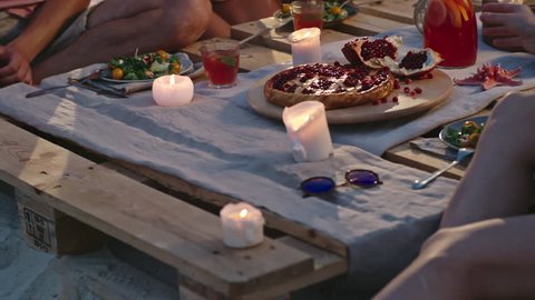 Group of young people having dinner at wooden beach table served with fruits, salads and tropical drinks and burning candles on it