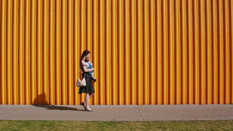 Young mother in a black-and-white dress carrying a baby boy along the yellow wall outside on a sunny day.
