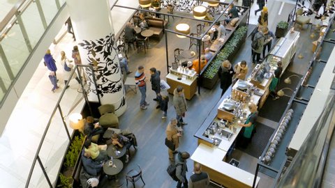 Buyers in the cafe choose food and drinks on display and talking to the cashier. Around the tables sit visitors. The view from the top