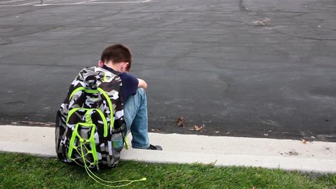 A sad, lonely boy sits on a curb at school with his head on his knees.