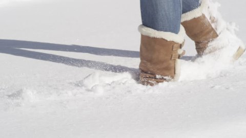 SLOW MOTION CLOSE UP: Unrecognizable female in jeans and warm winter boots walking in smooth deep snow blanket. Woman in sheepskin boots wading through fresh snow on a beautiful sunny Christmas day