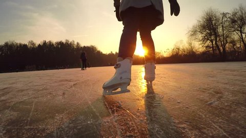 CLOSE UP, LOW ANGLE VIEW: Happy woman iceskating fast on frozen pond in local park at golden sunset on magical Christmas evening. People on ice skates enjoying winter activities in nature, having fun