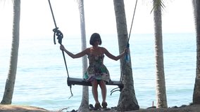 Young beautiful woman is having fun on a swing underneath a tree in nature, near the ocean. Very beautiful romantic ocean view, alone lady and swing. Bali island, Indonesia. Asian plants and palms
