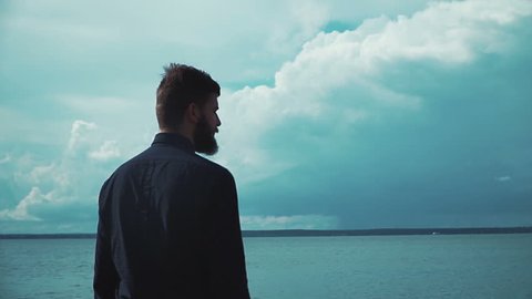 Young stylish man with a beard smokes an electronic cigarette in the background of the sea and clouds epic. Modern fashion vaping