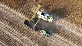 Harvester unloads sunflowers seed into farm vehicle truck HD video aerial top view in field. Harvest combine agriculture machine