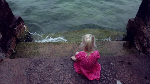 CINEMAGRAPH - Cute little girl sits on old stone dock at sea shore. Motion photo seamless loop స్టాక్ వీడియో