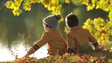 Two children, boys, sitting on the edge of a lake on a sunny autumn afternoon, watching swans swimming in the lake, beautiful autumn colors