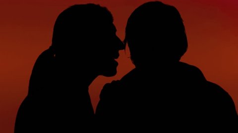 A woman whispering a secret in the ear of a man. Silhouette shot, red background.
