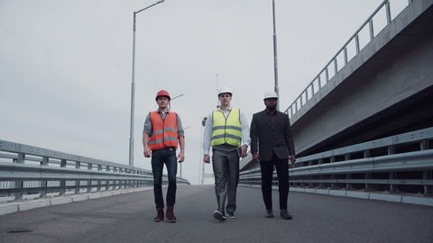 4K shot of Diverse group of three handsome male construction engineers with serious expressions walking on highway ramp. Wide angle
