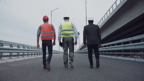 4K shot of Diverse group of three male construction engineers in white hard hats with reflective jackets walking on highway ramp. Rear view on road along bridge.