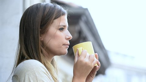1080 Young depressed woman on her 30s looking sad and thoughtful drinking tea or coffee cup at apartment balcony in female depression concept