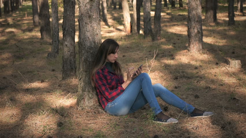 Young woman typing sms while sitting under pine tree