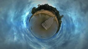 Tiny Little Planet 360 Degree Timelapse. Man, Backpacker is Walking by Coastline, Sandy Bank at Wooden Pier. Tourist is Traveling in Warm Autumn Day. Cloudy Sky. Fresh Green Trees Are Around, Park or