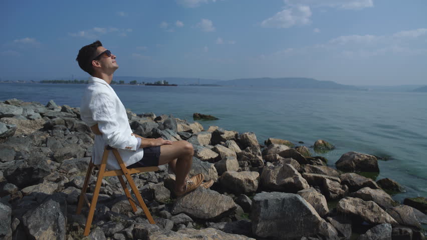 man in a white jacket sitting on a chair on the riverbank and enjoying the view