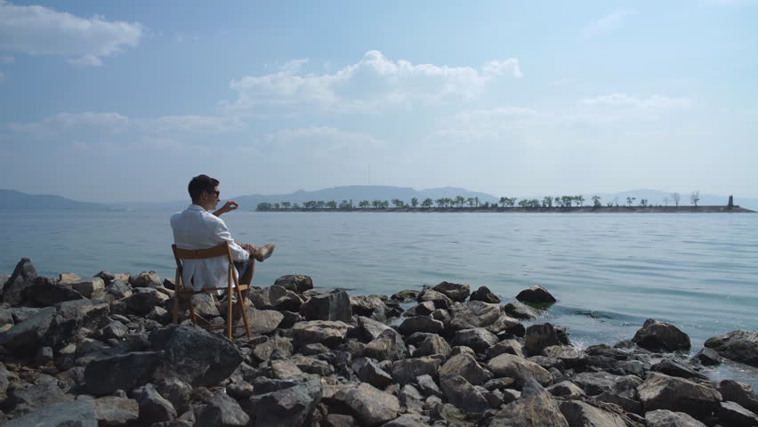 man in a white jacket sitting on a chair on the riverbank and enjoying the view