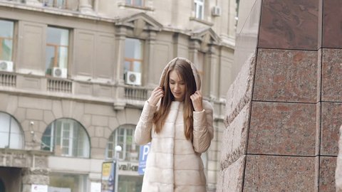 Stylish woman in furry coat passes, smiles and wears hood on the street. Slowly