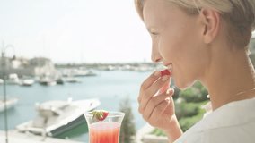 SLOW MOTION CLOSE UP DOF: Cheerful adult Caucasian woman on vacation eating sweet strawberry and drinking cocktail on hotel terrace, enjoying the view of sailboat harbor on amazing sunny summer day - Powered by Shutterstock - Get 15% off with code: PIKWIZARD15