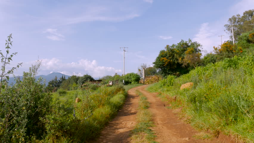 A man in a hat walks away from the camera down a dirt road and sits on a rock and puts his knee up