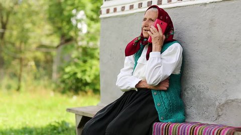 Senior woman sits on bench and talks on mobile phone. Old woman sits on bench near his house and speaks on cell phone. Ukrainian old woman with red smartphone. Female with smartphone. Roar of laughter