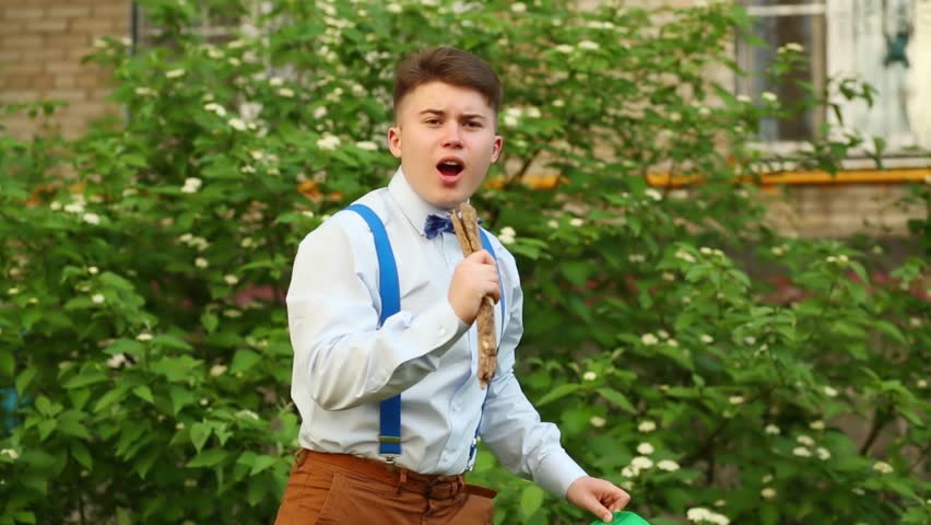 Boy in suspenders and necktie, with stick in hand pretending to sing near green bushes.