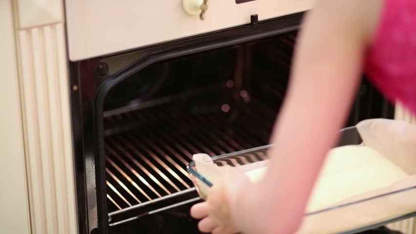 Little girl is putting glass pan with batter to the conventional oven.