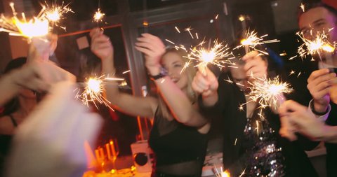Handheld shot of carefree young men and women with glasses of champagne waving sparklers around, dancing and posing for camera in dark night club
