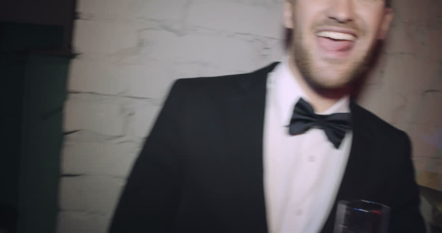 Handheld shot of tipsy stylish young man in suit leaning on brick wall in night club drinking champagne and laughing to camera