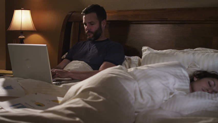 couple in bed. man works on laptop computer in bed while a woman sleeps.