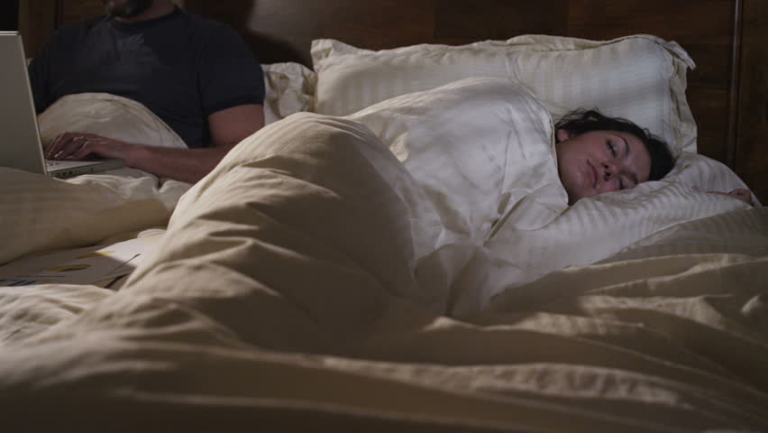 couple in bed. man works on laptop computer in bed while a woman sleeps.