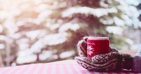 Hands in Knitted Mittens holding Steaming Cup of Hot Coffee or Tea on Snowy Winter Morning Outdoors. 4K DCi SLOW MOTION 120 fps. Woman holds Cozy Festive Red Mug with a Warm Drink on Christmas Morning