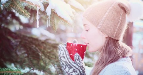 Portrait of a Smiling Woman with Cup of Hot Tea or Coffee Enjoying Cozy Snowy Winter Morning Outdoors. 4K DCi SLOW MOTION 120 fps. Beautiful Girl with a Steaming Mug by Snow Covered Christmas Tree.