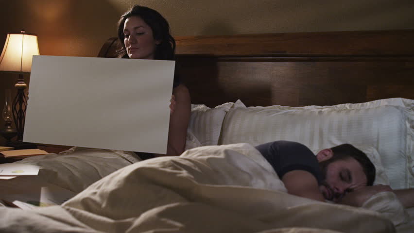 couple in bed. woman holds blank sign while a man sleeps.