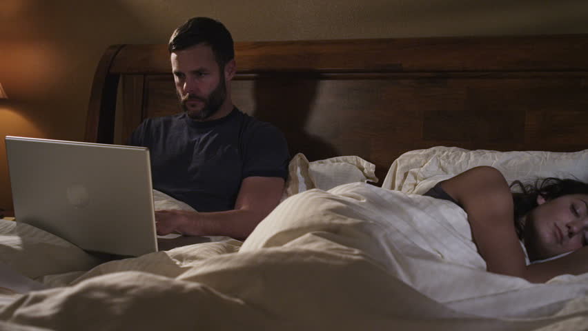 couple in bed. man works on laptop computer in bed while a woman sleeps.