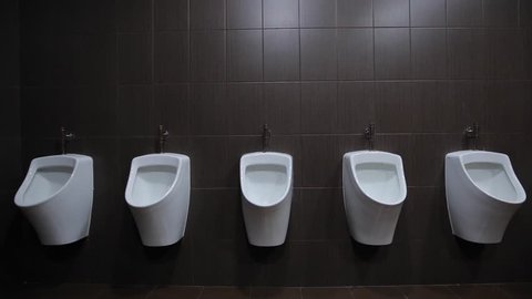 Urinals are fixed on a white wall, gray tiles on the walls, brown floor tiles, taps for urinals silver metal. survey conducted indoors under fluorescent light bulbs, front view. wide angle, static