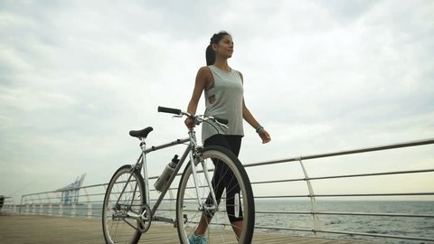 Young active brunette holding bycicle in her hand, while posing on camera.